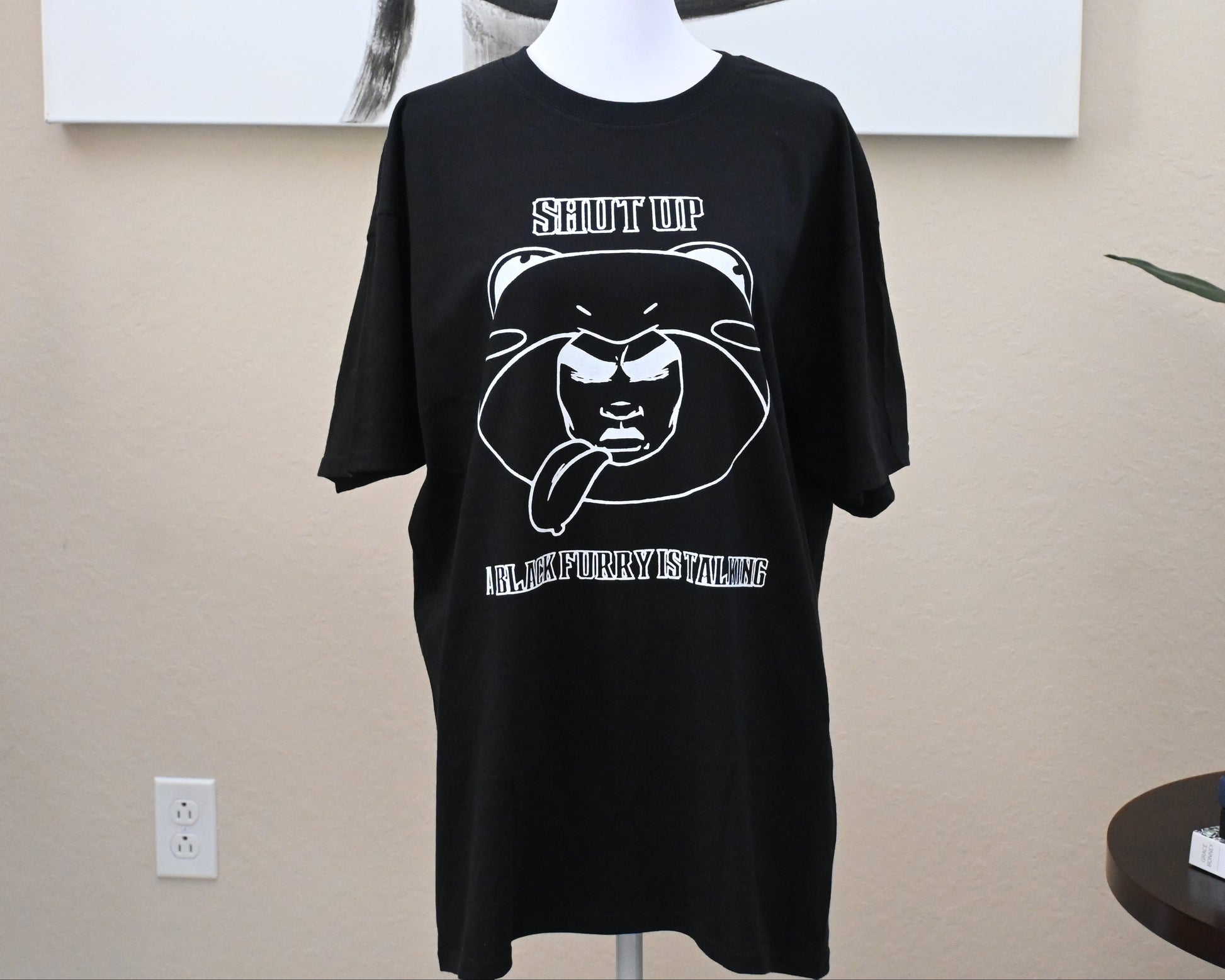 Black t-shirt with graphic design on a mannequin against a beige wall.