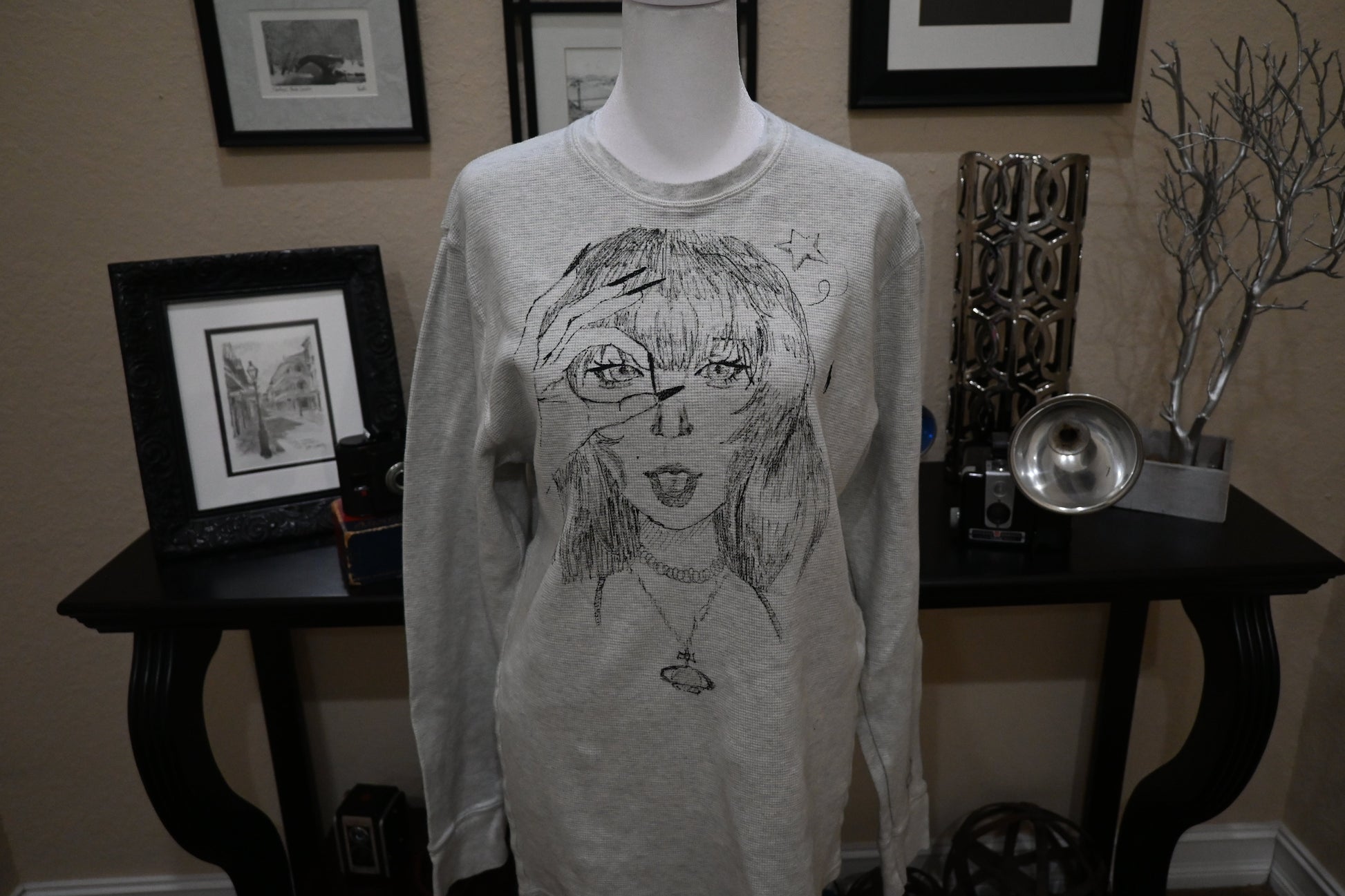 Mannequin wearing a long-sleeve shirt with a graphic design in a room with framed pictures on the wall.