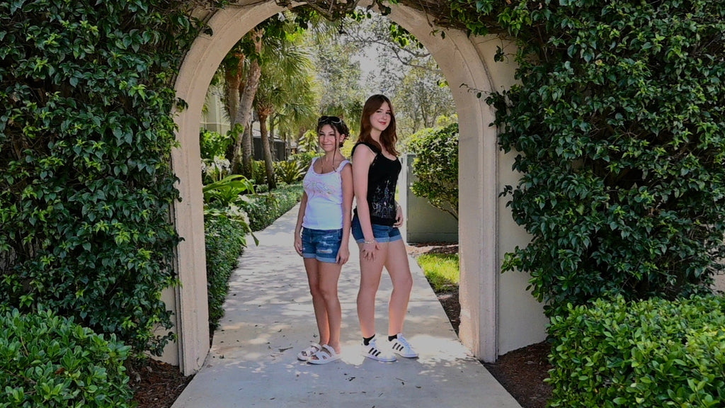 Two girls wearing hand painted lacy tank tops. One design is a green dragonfly with pink designs, and the other is a black shirt with a white cross and church.
